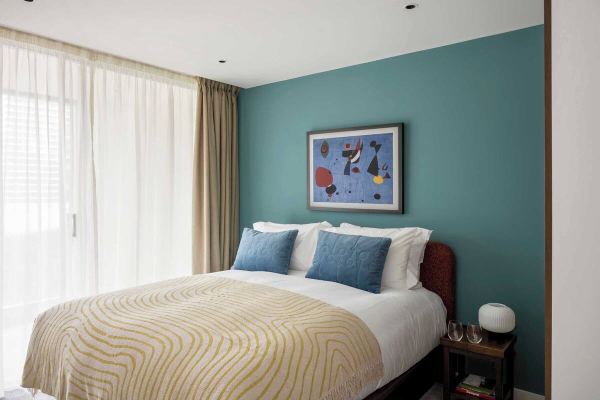 A neatly made bed with a patterned blanket, a bedside table with a lamp and glasses, and a painting on the wall.