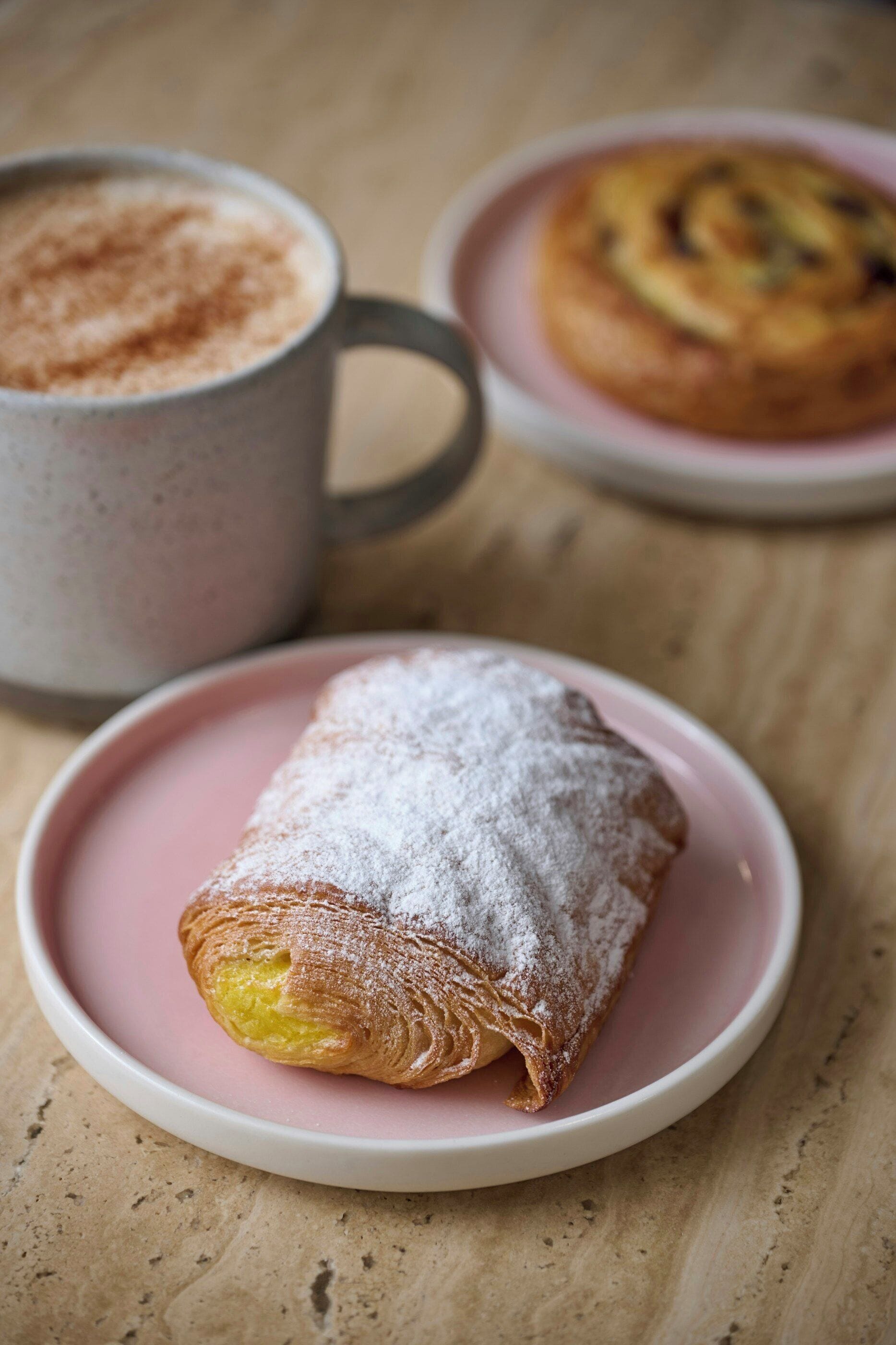 A pastry on a pink plate, a mug, and another plate with a pastry in the background.