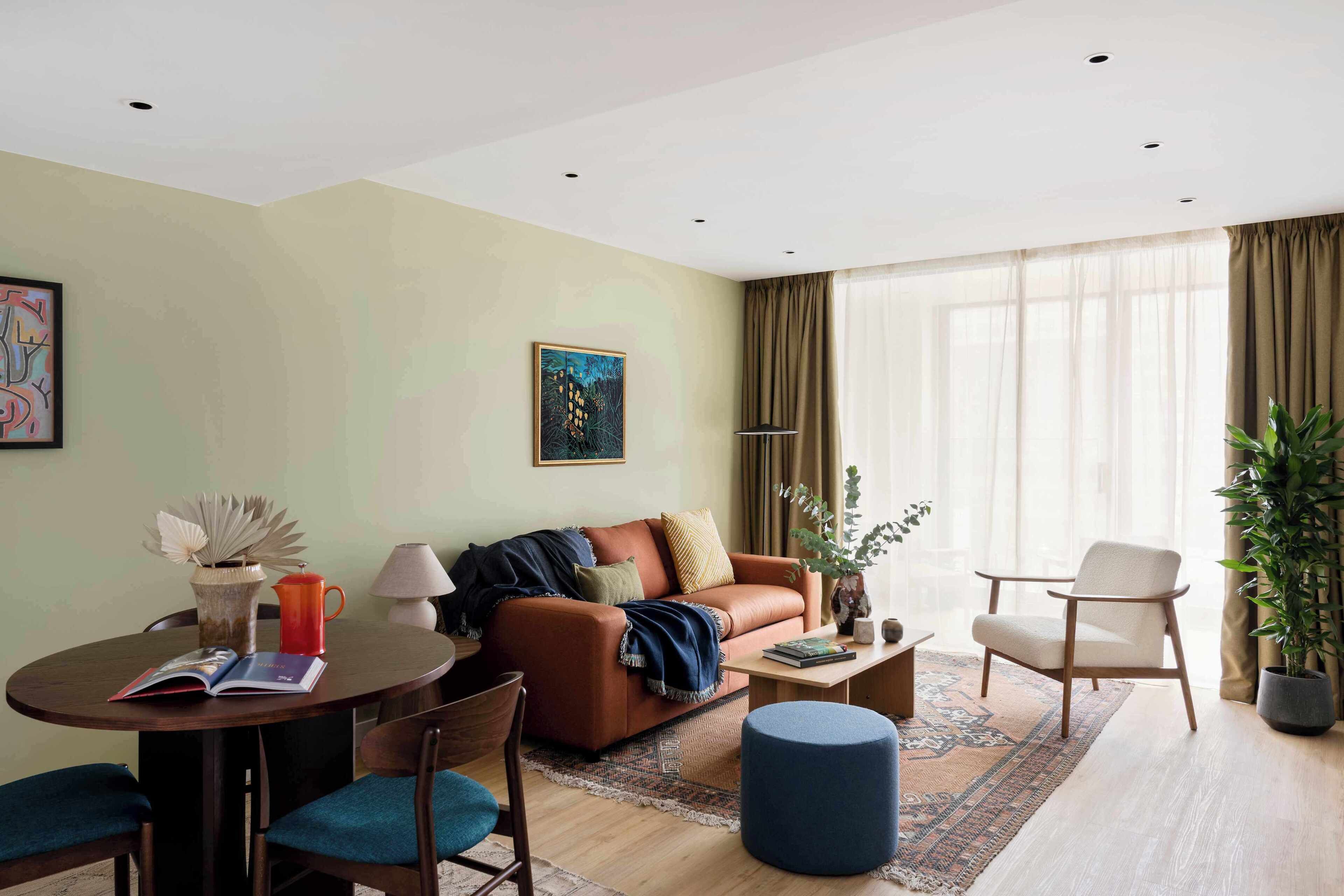 A modern living room with a brown leather sofa, a round coffee table, and a blue ottoman. There is a dining table with chairs and a lamp.