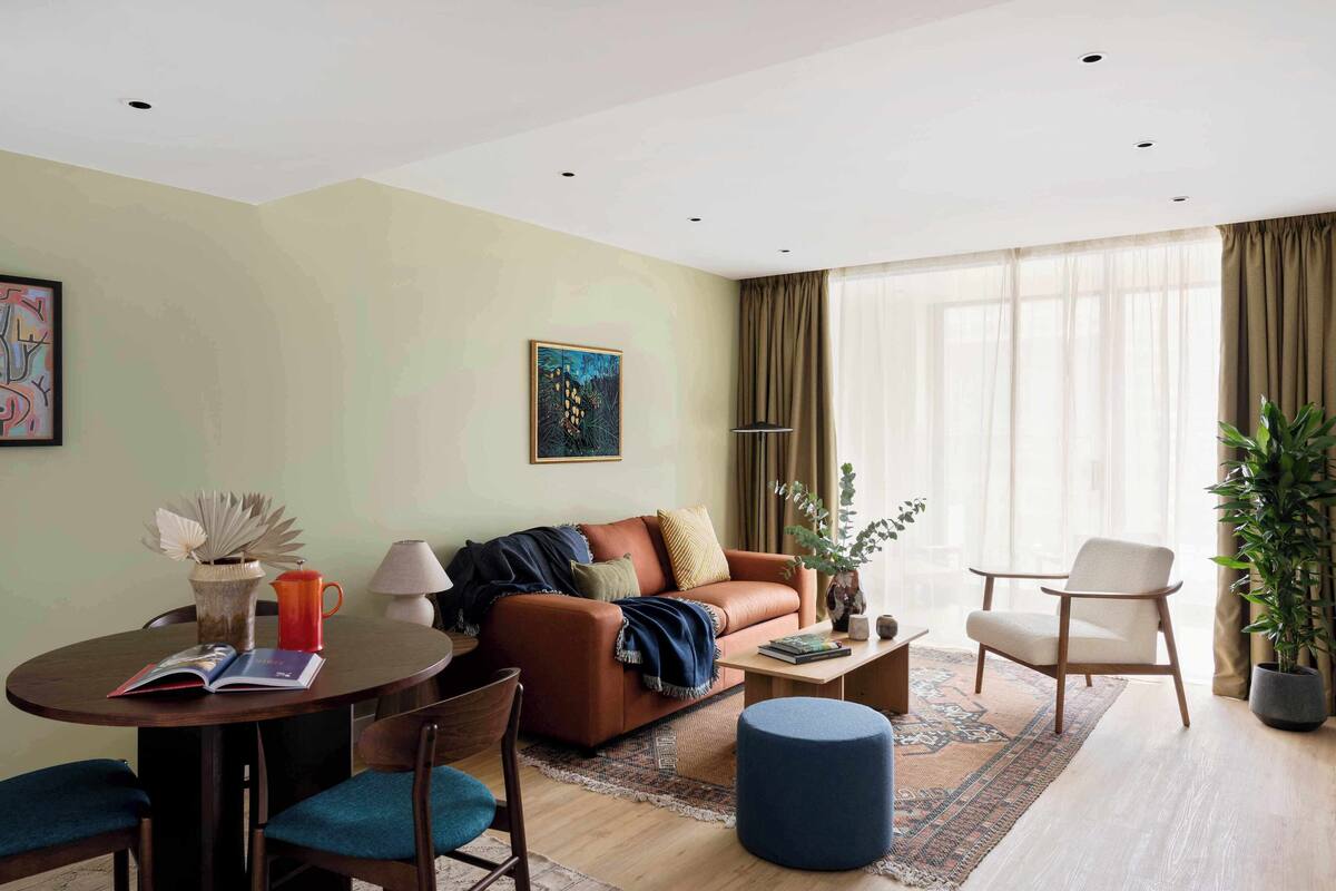 A modern living room with a brown leather sofa, a round coffee table, and a blue ottoman. There is a dining table with chairs and a lamp.