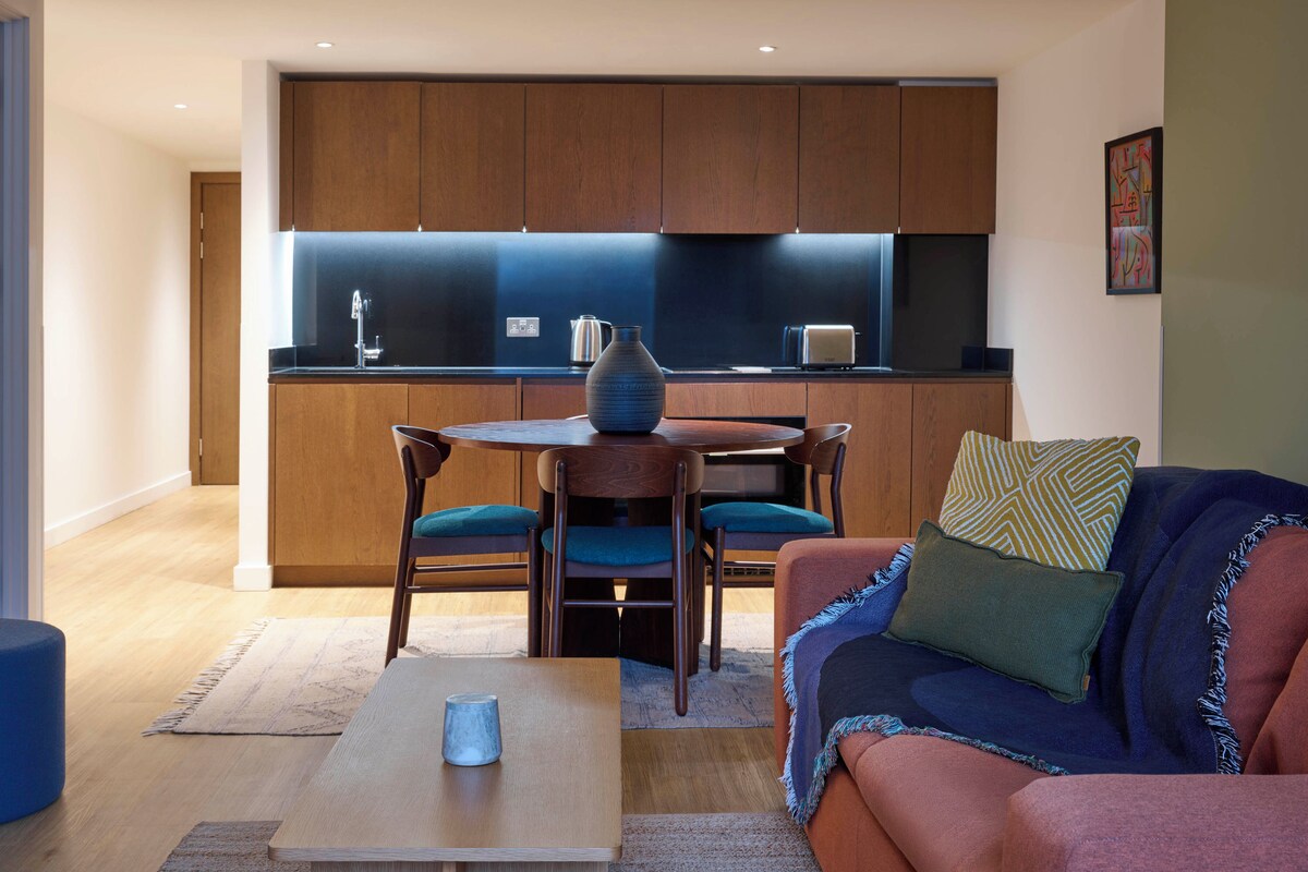 A modern kitchen with wooden cabinets, a dining area with a round table and chairs, and a cozy living space with a sofa and cushions.