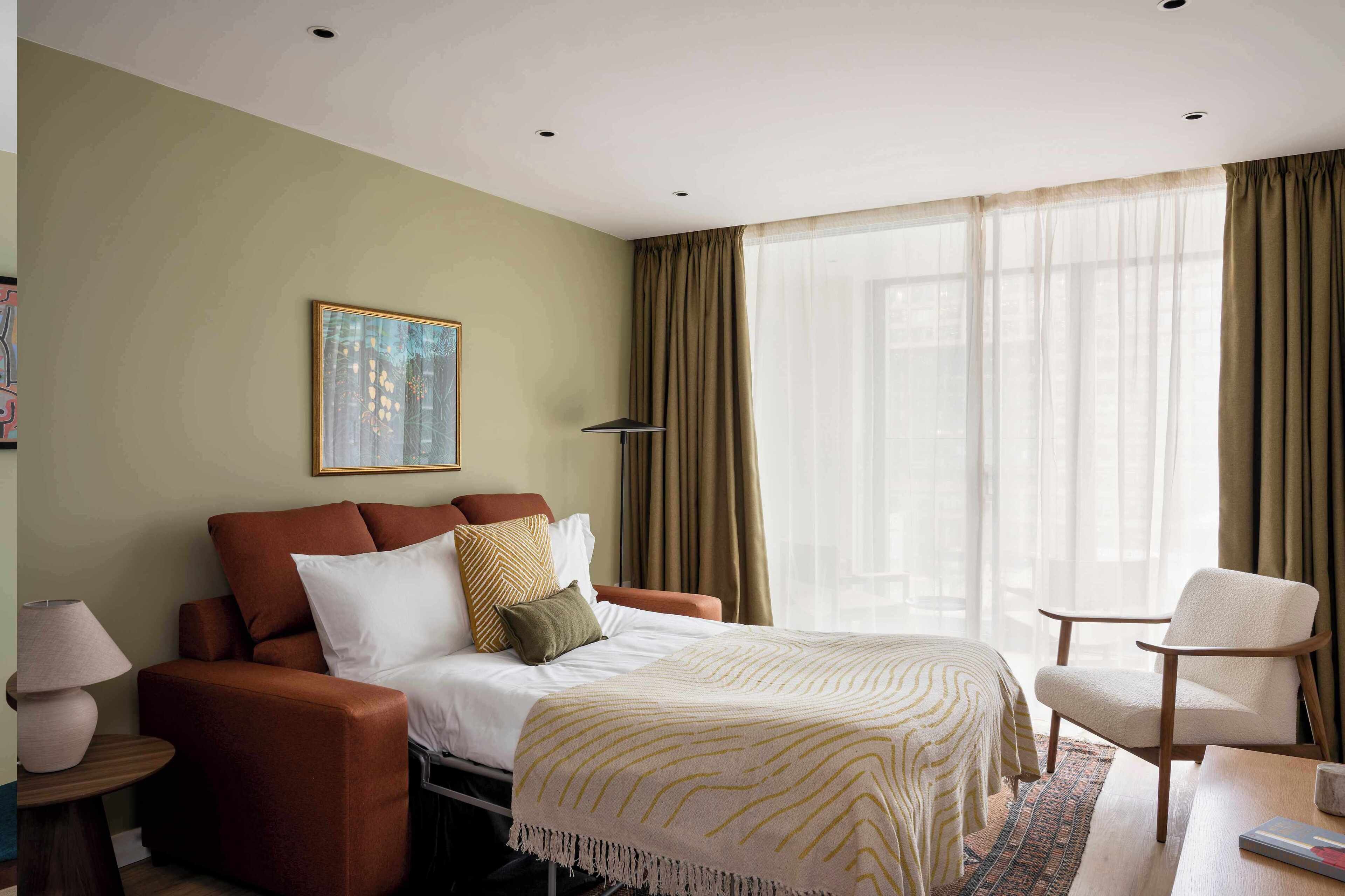 A hotel room with a bed, a sofa, a chair, a lamp, and a painting on the wall.