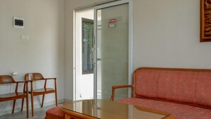 Ein Zimmer mit Glastür, die auf einen Balkon führt, einem Holztisch, zwei Holzstühlen und einem gemusterten Sofa.