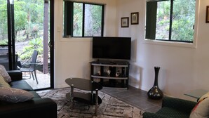 Een woonkamer met een bank, een salontafel, een flatscreen televisie op een meubelstuk, een vaas en uitzicht op de natuur door grote ramen.