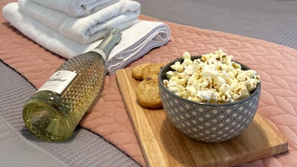 En snyggt bäddad säng med rosa kuddar, en hög med vita handdukar, en flaska olivolja, en kaka och en skål med popcorn på en rosa, vadderad yta.