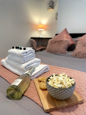 En snyggt bäddad säng med rosa kuddar, en hög med vita handdukar, en flaska olivolja, en kaka och en skål med popcorn på en rosa, vadderad yta.