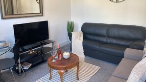 Ett vardagsrum med en svart lädersoffa, ett träbord med en flaska och glas på, en platt-TV på ett stativ, en stor väggklocka och en spegel ovanför TV:n.