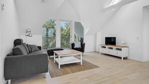 Un salon spacieux avec un plafond haut, une grande fenêtre donnant sur les arbres, un téléviseur à écran plat et un meuble de rangement blanc pour les appareils multimédias.