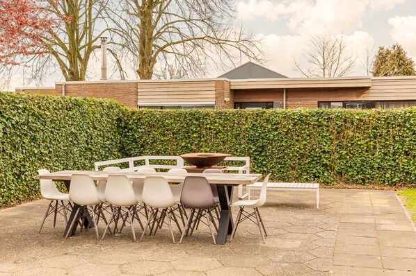 Un patio al aire libre con una mesa, sillas, un seto y un edificio al fondo.