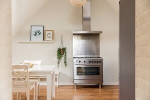 Una cocina con estufa de acero inoxidable, una mesa de comedor blanca con sillas y una planta colgante.