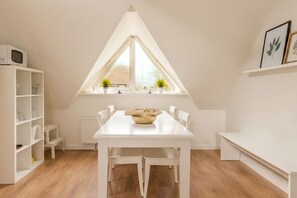 Un comedor con una mesa y sillas blancas, un estante blanco y una ventana con plantas.