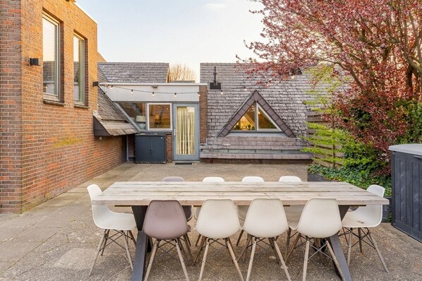 Un patio con una mesa y sillas de madera, un muro de ladrillo y un edificio con techo inclinado.