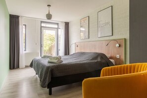Une chambre à coucher comprenant un lit, une table de chevet, une chaise, une fenêtre avec des rideaux et un mur orné d’un tableau encadré.