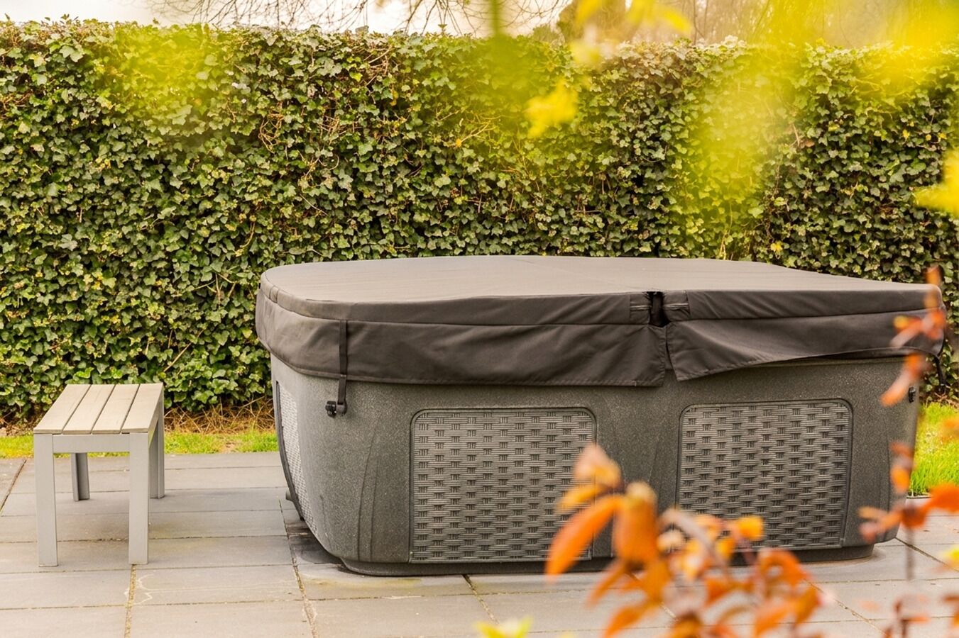 A grey outdoor heater with a mesh front panel, a small wooden stool, and a hedge in the background.