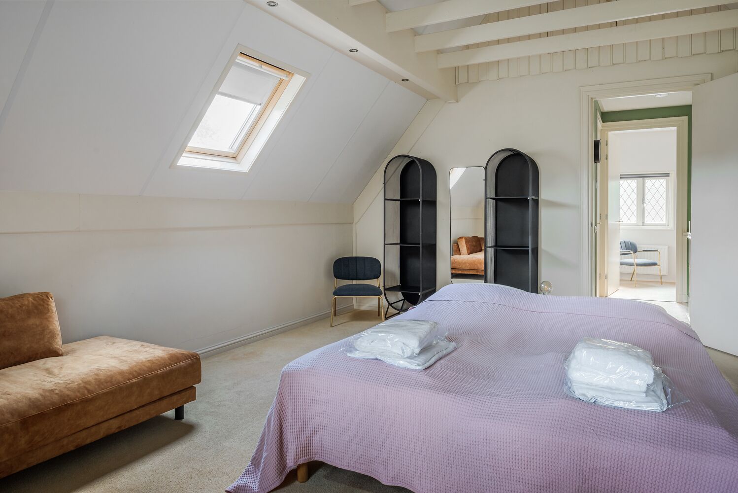 Quarto com teto inclinado, uma cama com colcha roxa, sofá castanho e dois móveis de armazenamento pretos.