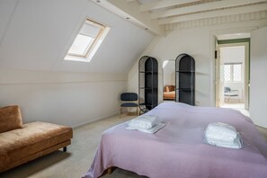 Un dormitorio con techo inclinado, una cama con edredón morado, un sofá marrón y dos unidades de almacenamiento negras.