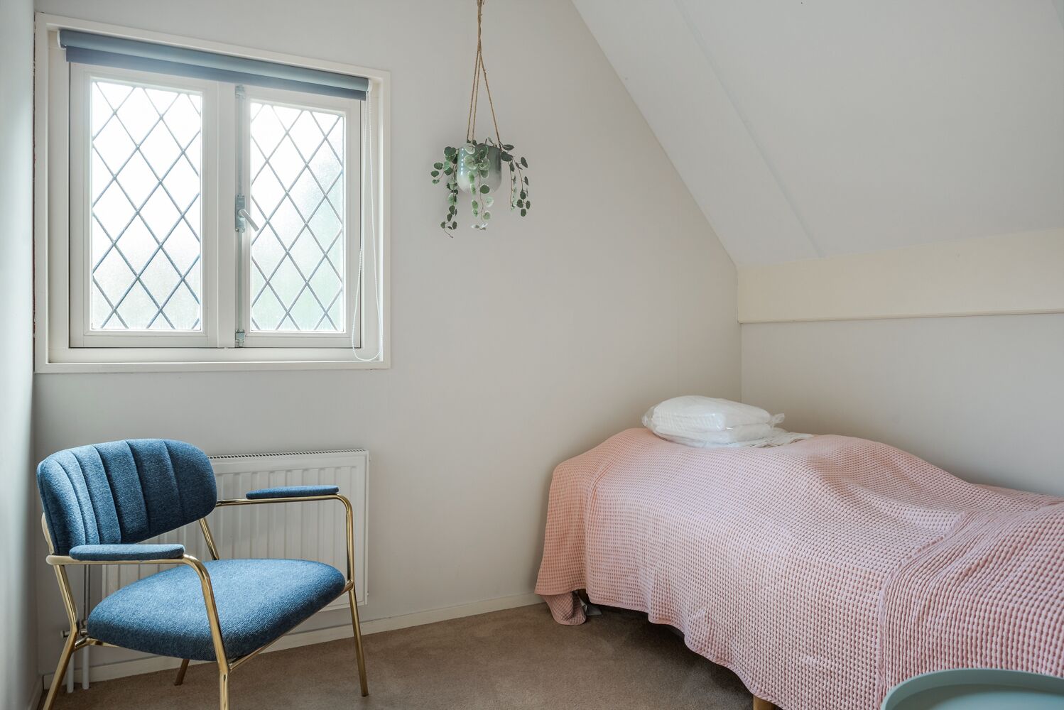 A bedroom with a bed, a chair, a radiator, and a window with a plant hanging from the ceiling.