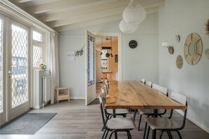 Un comedor espacioso con una larga mesa de madera y sillas modernas, un techo blanco con vigas a la vista y una vista a un balcón exterior a través de un ventanal.