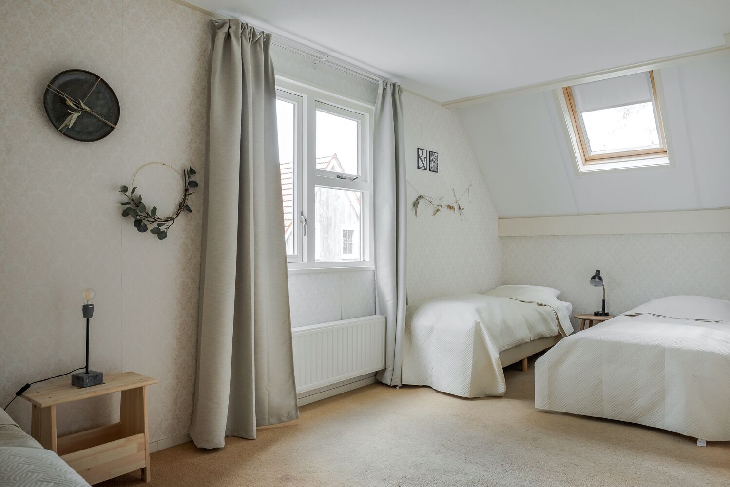 A bedroom with two beds, a bedside table, a lamp, a clock, and a window with curtains.