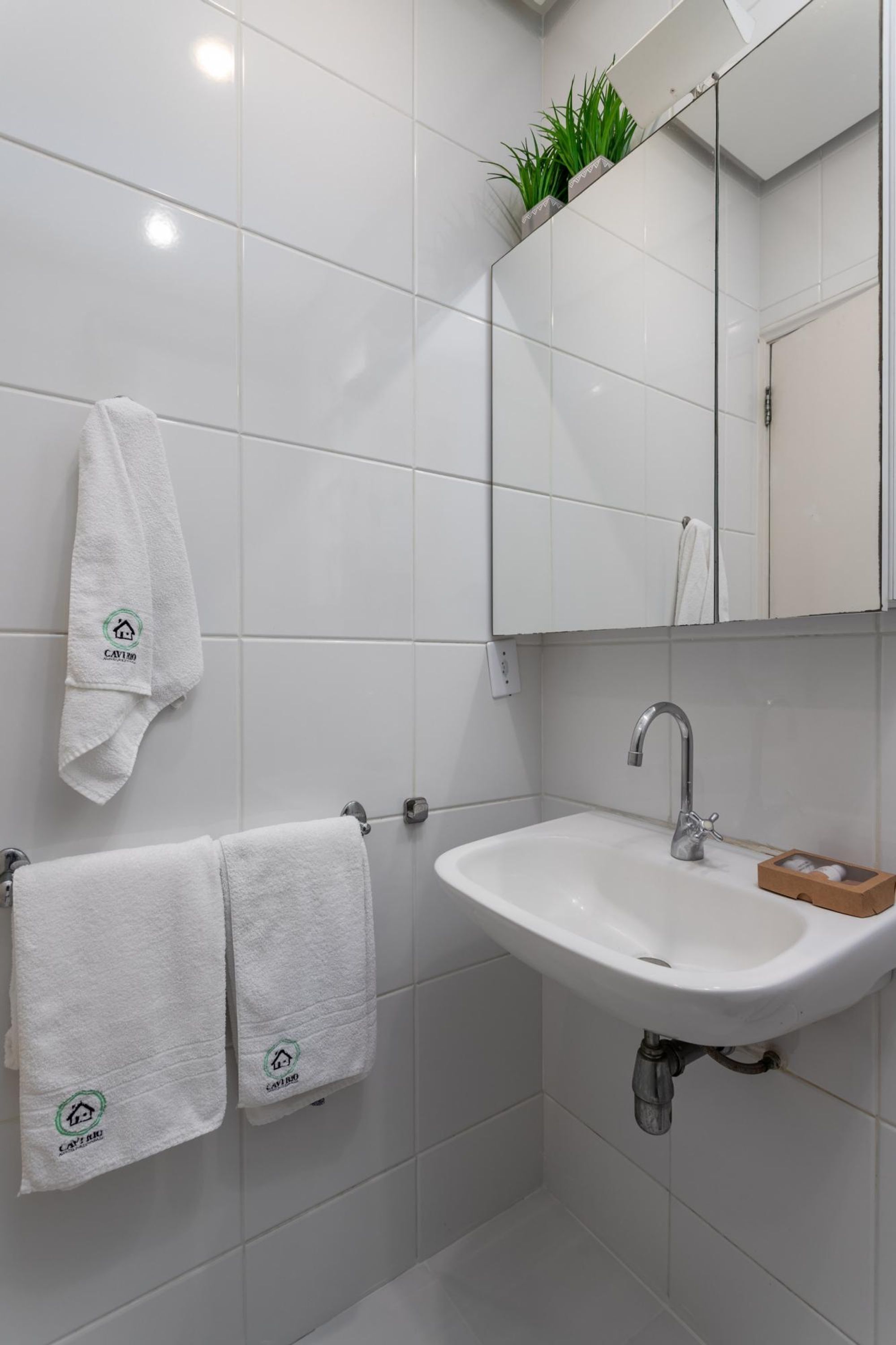 Un baño con azulejos blancos, un lavamanos, un espejo y dos toallas con un logotipo.