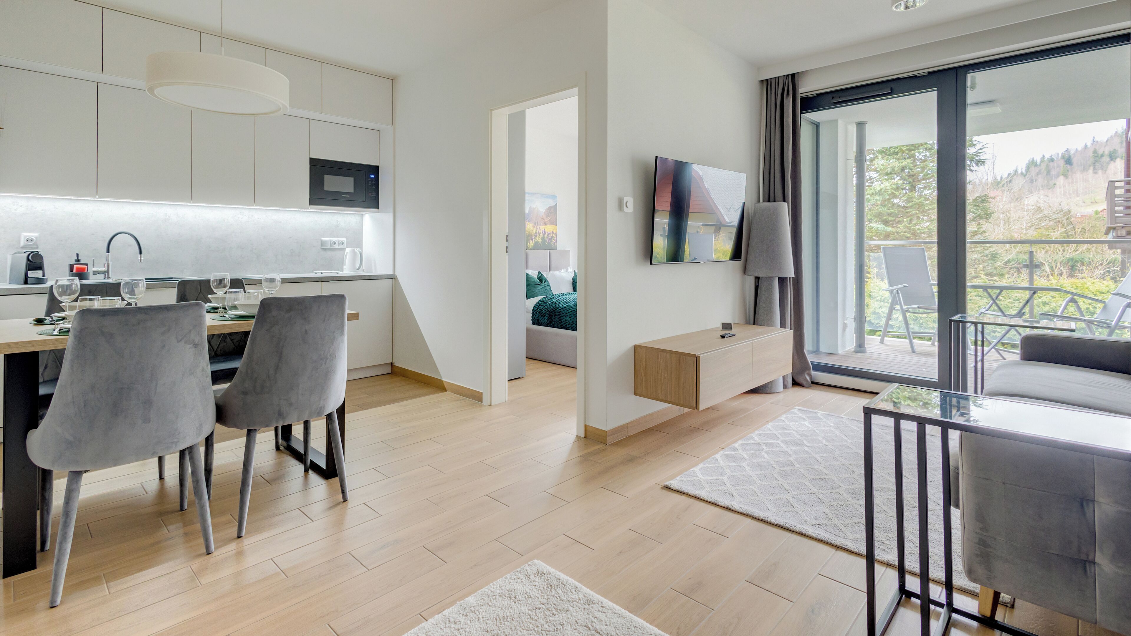 Una cocina moderna con microondas, comedor con sillas, un sofá y un balcón con vista a árboles.