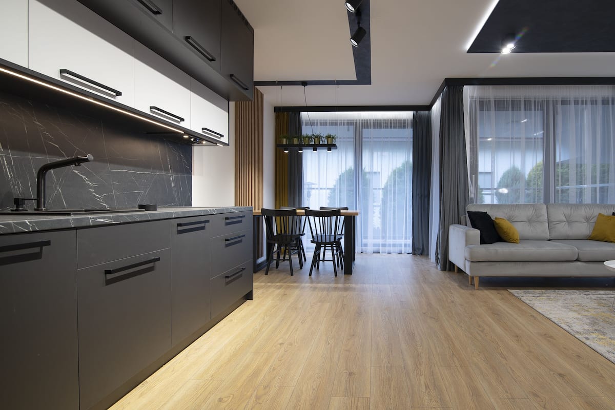 Een moderne keuken met donkere kasten en een marmeren aanrecht, een eethoek met een houten tafel en stoelen, en een zithoek met een bank en salontafel.