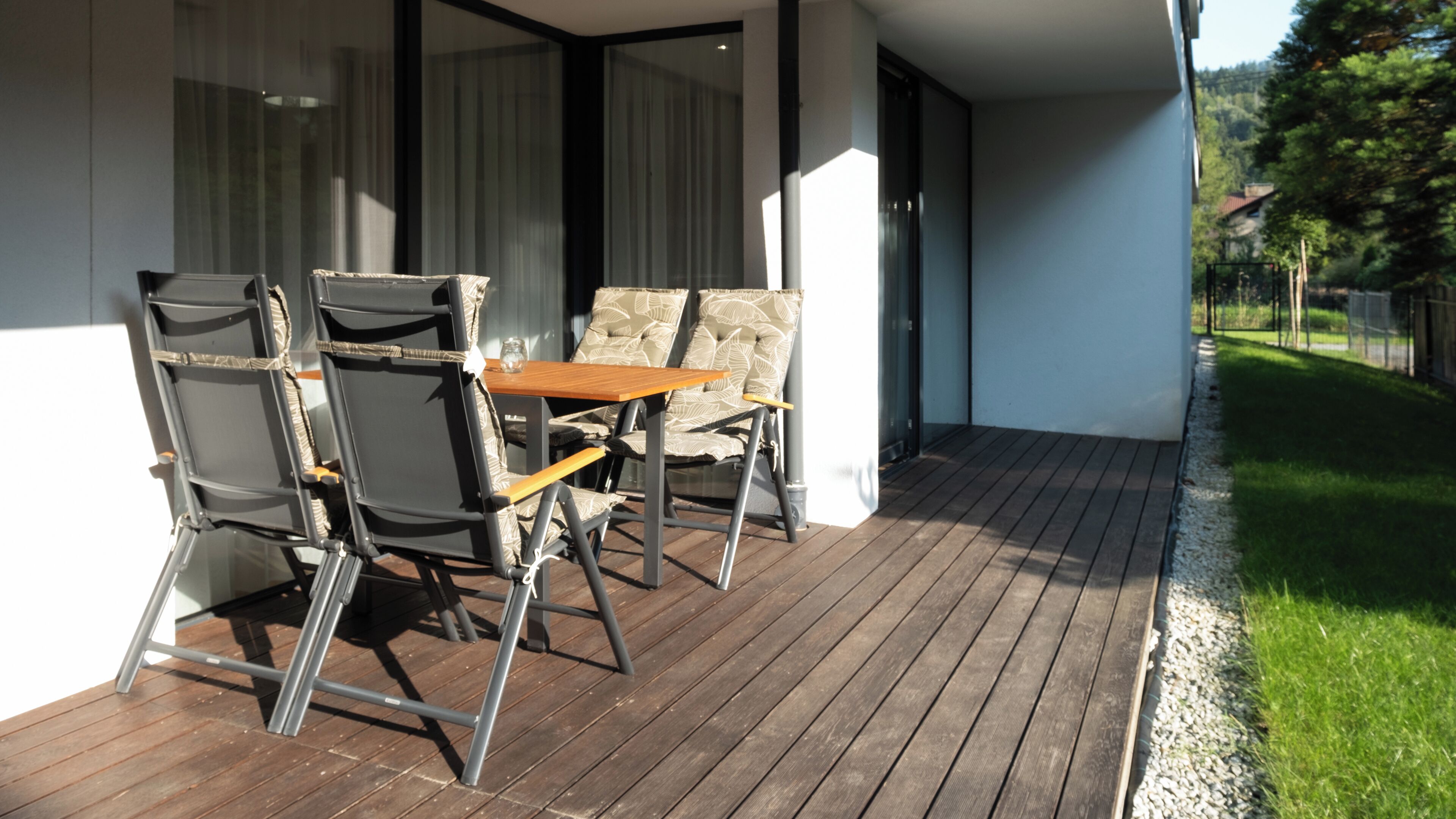 Een houten terras met een tafel en stoelen, een glazen deur naar een binnenruimte en een gazon met een hek.