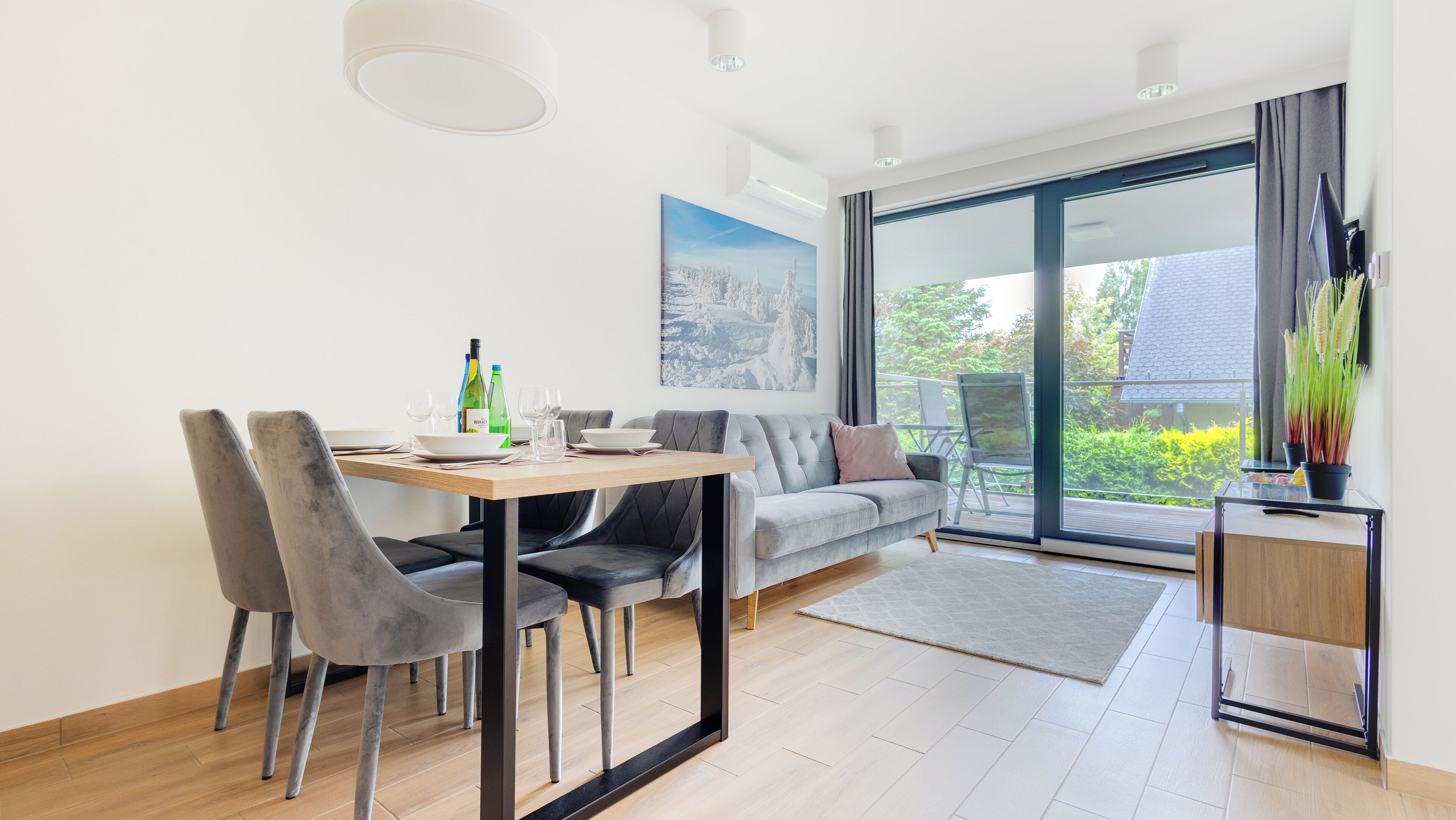 A modern living room with a dining area, a grey sofa, a glass table, and a large window offering a view of greenery.