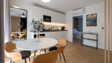 Una cocina moderna con armarios blancos, horno empotrado y cafetera. Hay una zona de comedor con una mesa redonda y sillas, y una pequeña planta sobre la mesa.