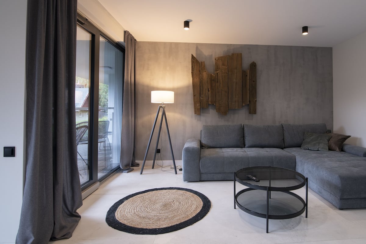 Een moderne woonkamer met een grijze hoekbank, een ronde salontafel, een vloerlamp en een groot raam met gordijnen.