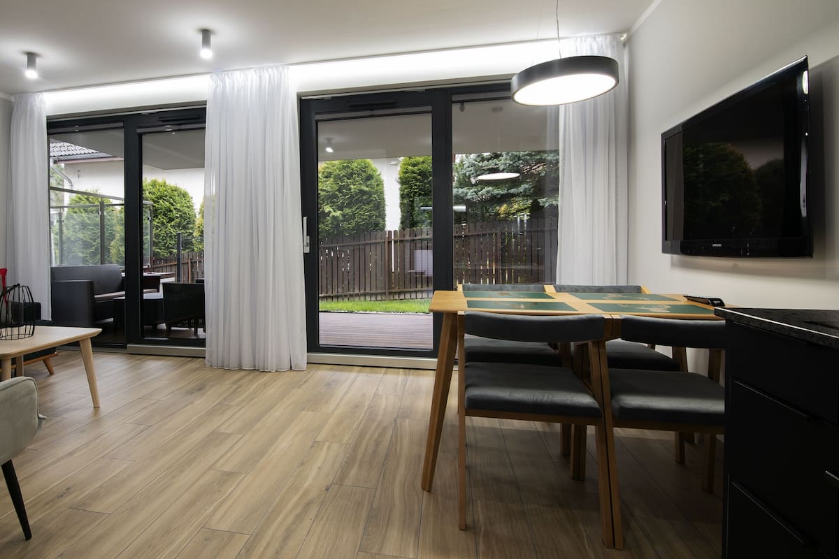 Een moderne woonkamer met een eethoek, grote schuifpuien naar een achtertuin, en een flatscreen televisie aan de muur.