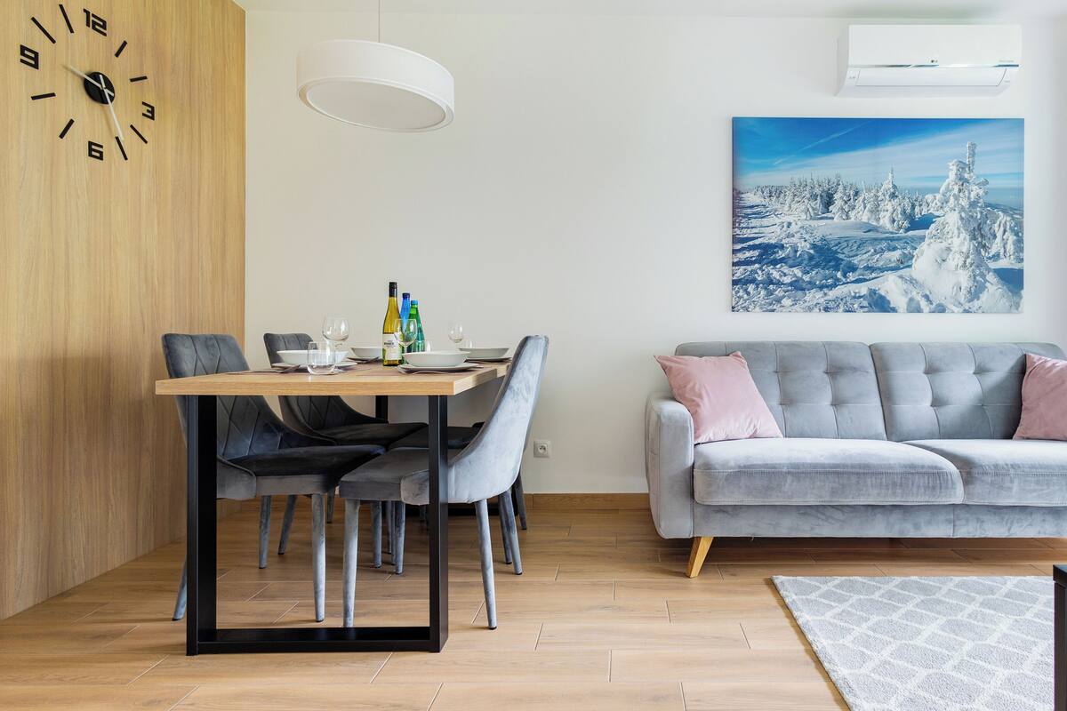 Een moderne woonkamer met een eetgedeelte, met een grijze bank, een houten eettafel en een groot raam met uitzicht op een besneeerd landschap.