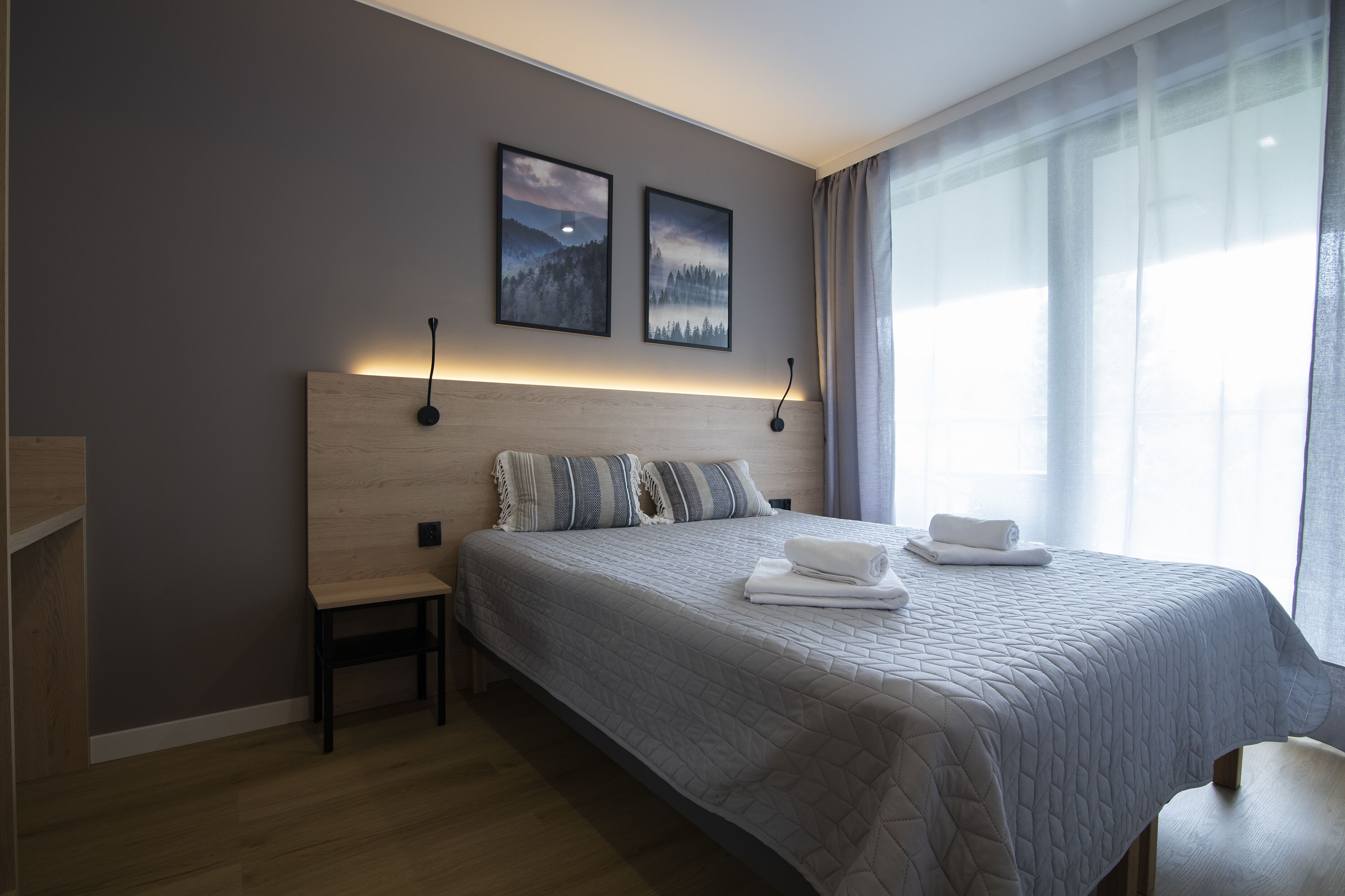 A modern hotel room with a large bed, a bedside table, a small chair, and two framed pictures on the wall.