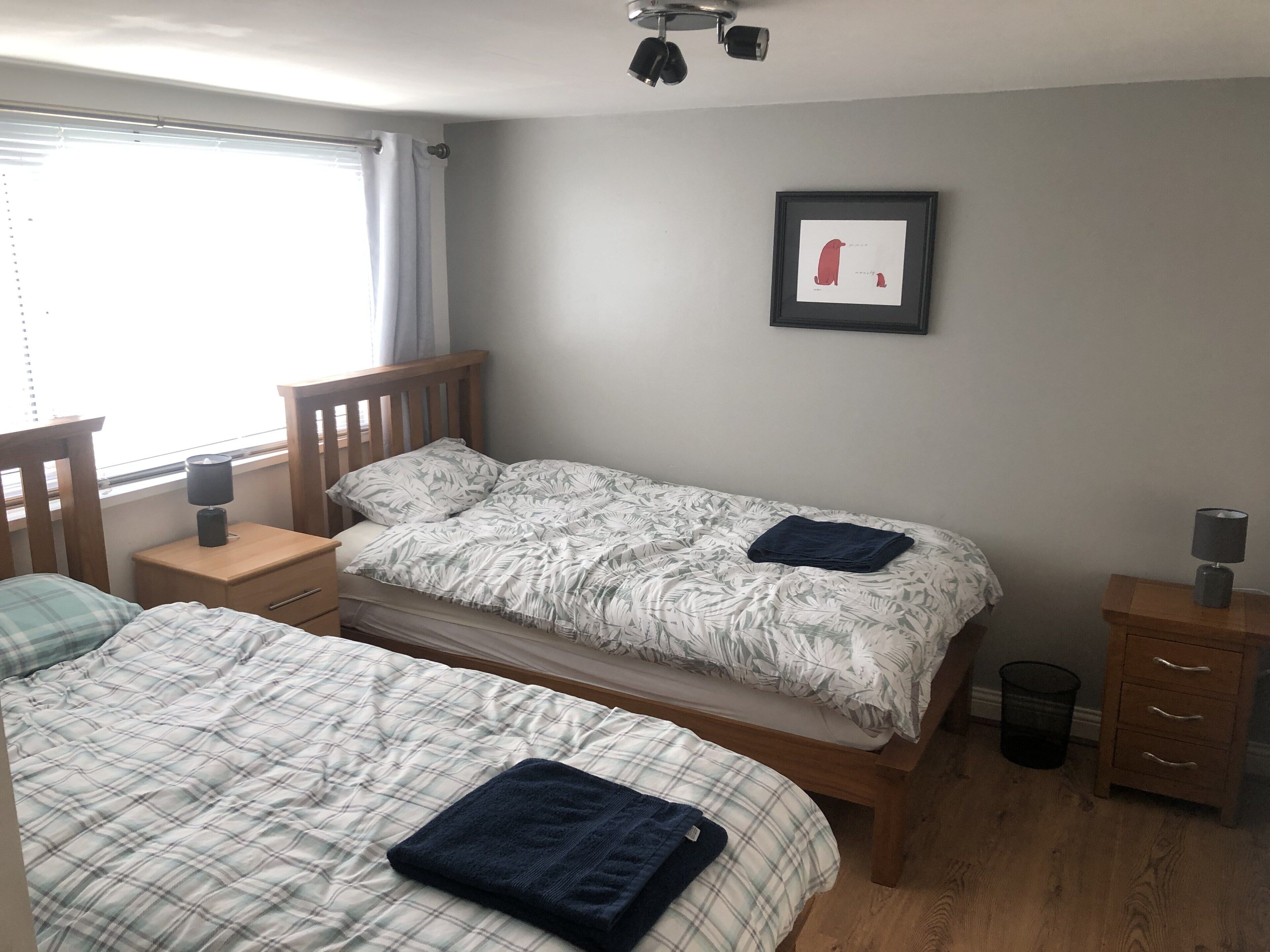 A bedroom with two beds, a small bedside table with a lamp, a framed picture on the wall, and a window with curtains.