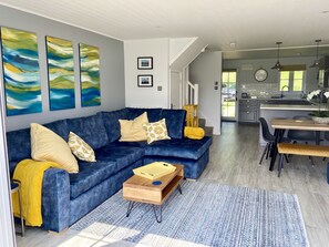 Un moderno salón con un sofá seccional azul, cojines amarillos y una mesa de centro de madera. Se observan pinturas abstractas en la pared, una cocina al fondo y un comedor con mesa y sillas de madera.
