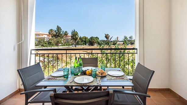 Un balcón con una mesa lista para dos, con vista a una playa con palmeras y edificios al fondo.
