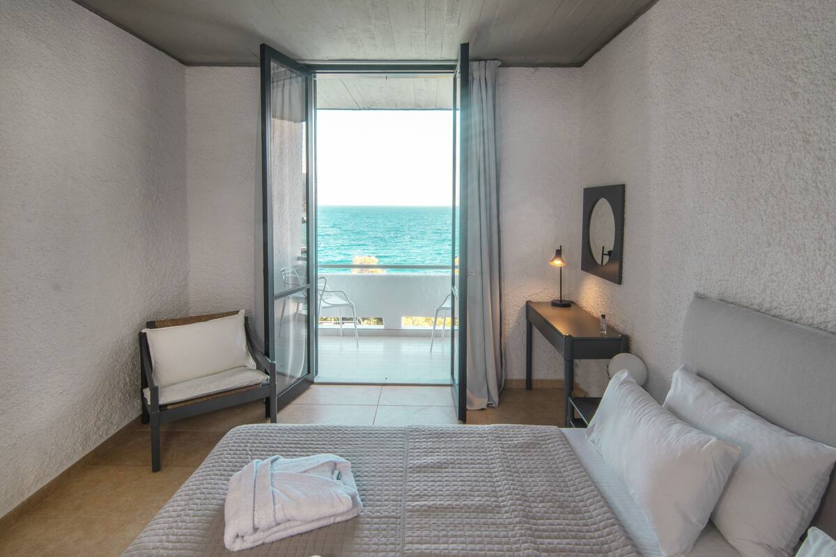 A bedroom with a bed, a chair, a table, a lamp, and a view of the sea through an open door.