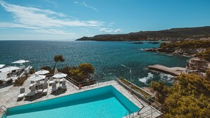 Una piscina con sillones de descanso y sombrillas, con vistas a una zona costera de aguas azules cristalinas y colinas lejanas.