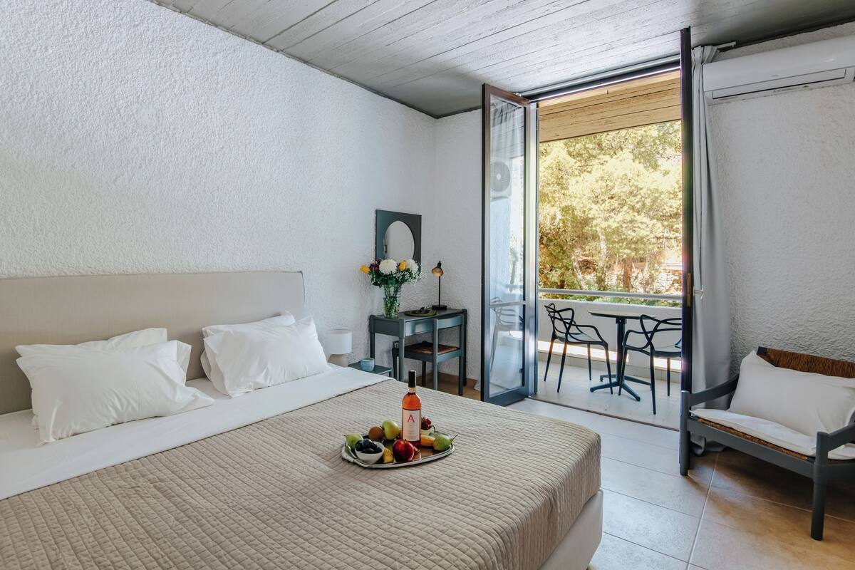 A bedroom with a bed, a bedside table with a lamp, a small table with a vase of flowers, and a balcony with chairs.
