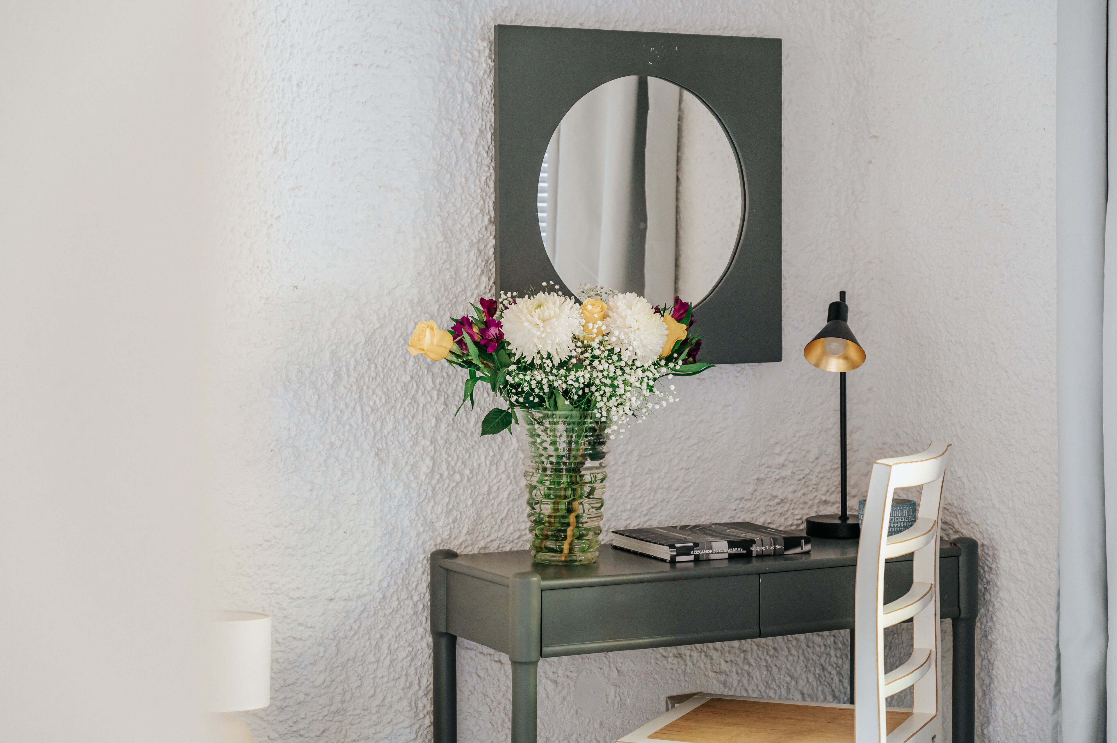 A desk with a mirror, a vase of flowers, a lamp, and a chair.