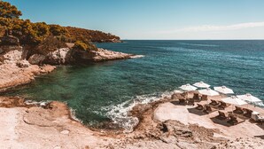 Una playa costera con aguas claras, terreno rocoso y varias sombrillas y sillas de playa blancas.