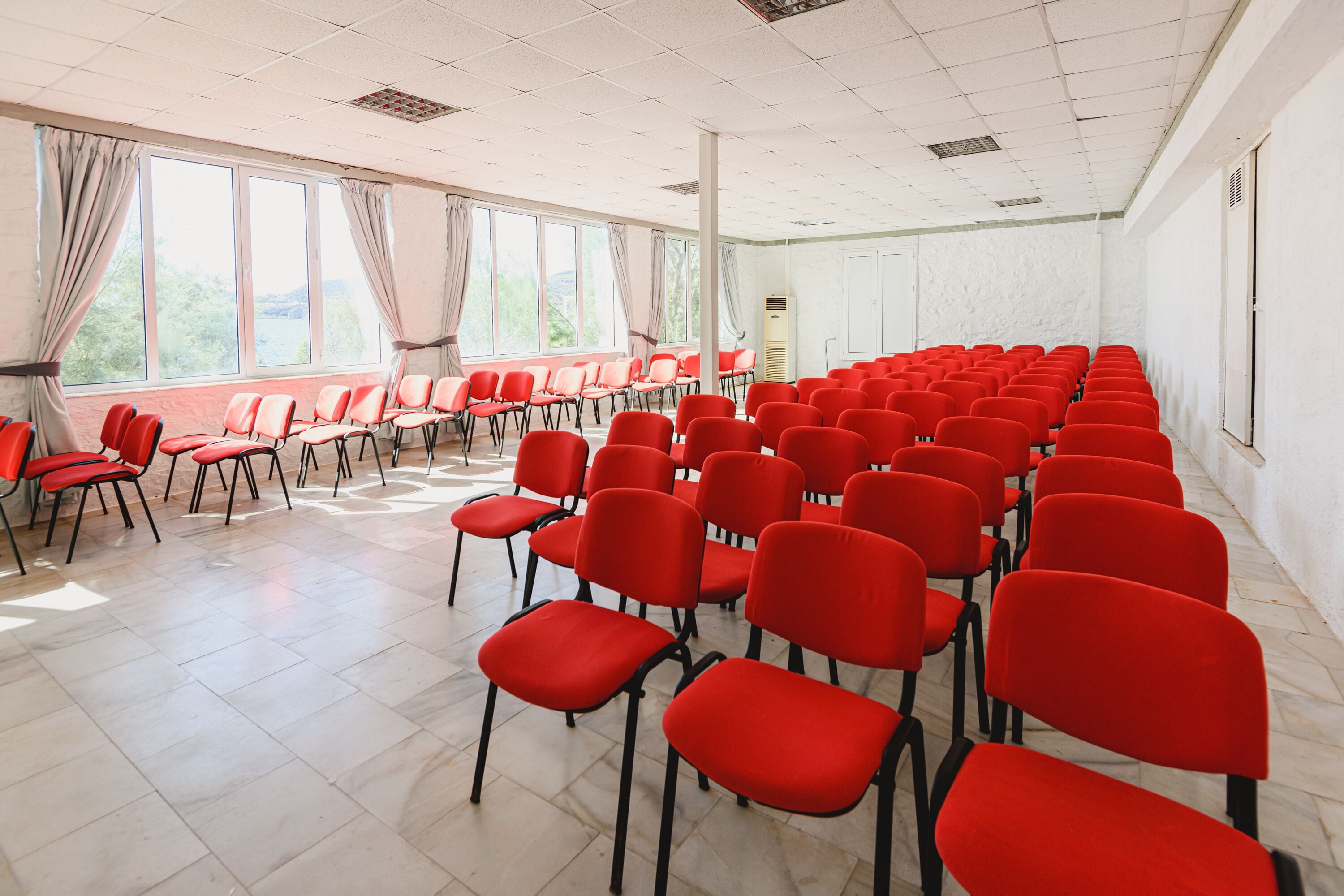 A conference room with rows of red chairs, large windows with curtains, and a white tiled floor.