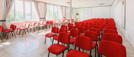 A conference room with rows of red chairs, large windows with curtains, and a white tiled floor.