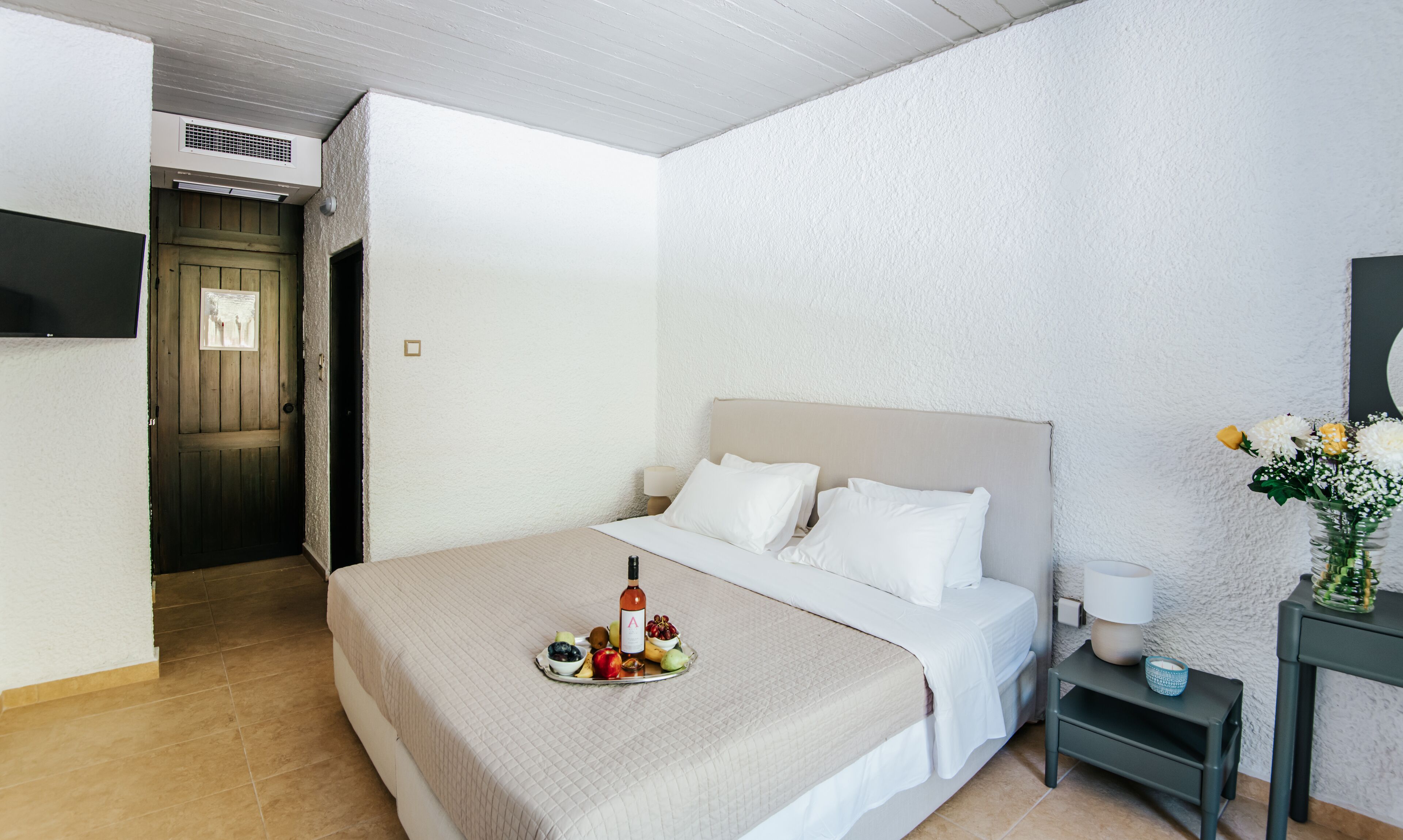 Ein ordentlich eingerichtetes Hotelzimmer mit Bett, Nachttisch und einem kleinen Tisch mit Flasche und Früchten.
