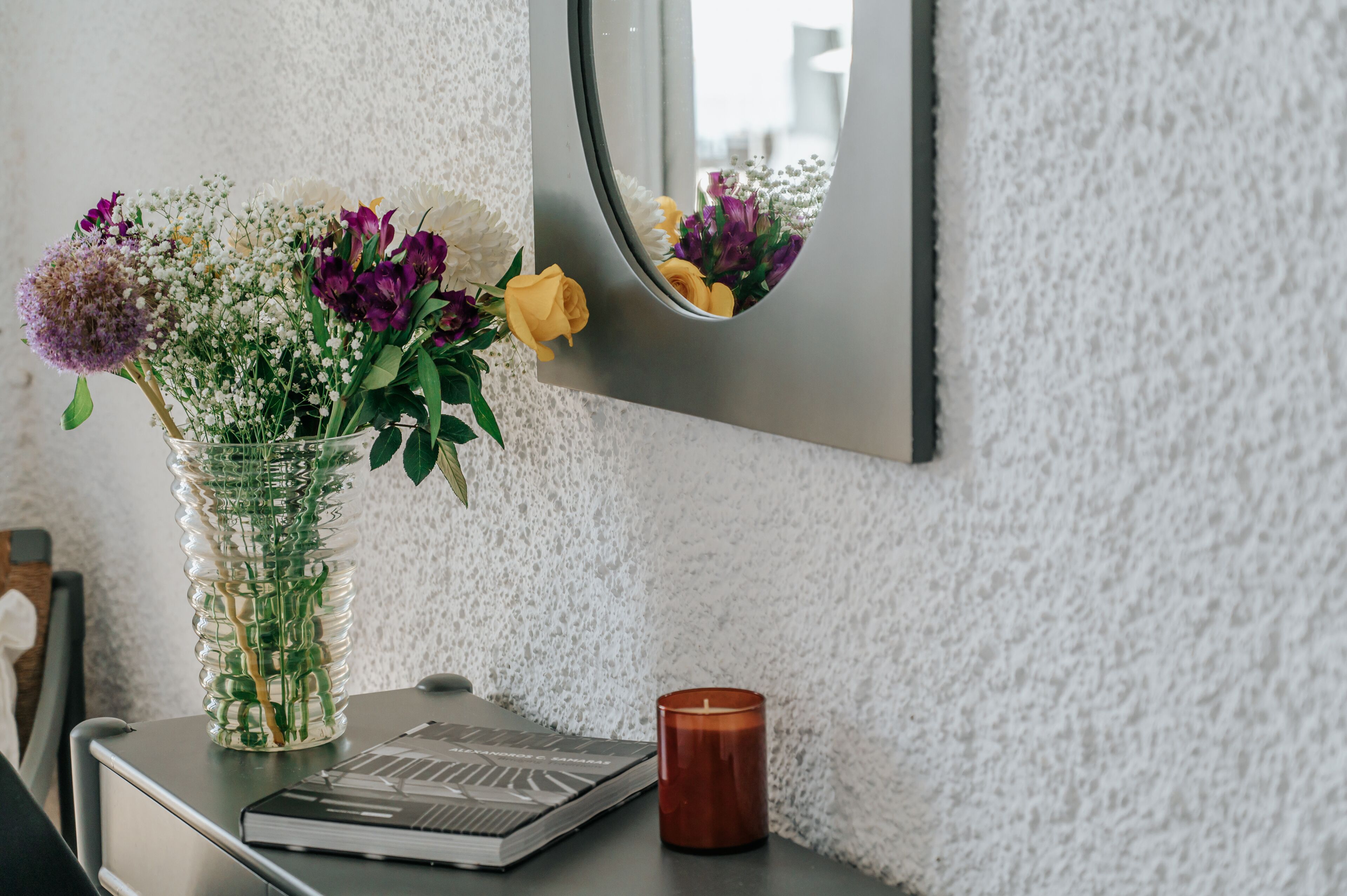 A vase with flowers on a table, a candle, and a mirror on a textured wall.