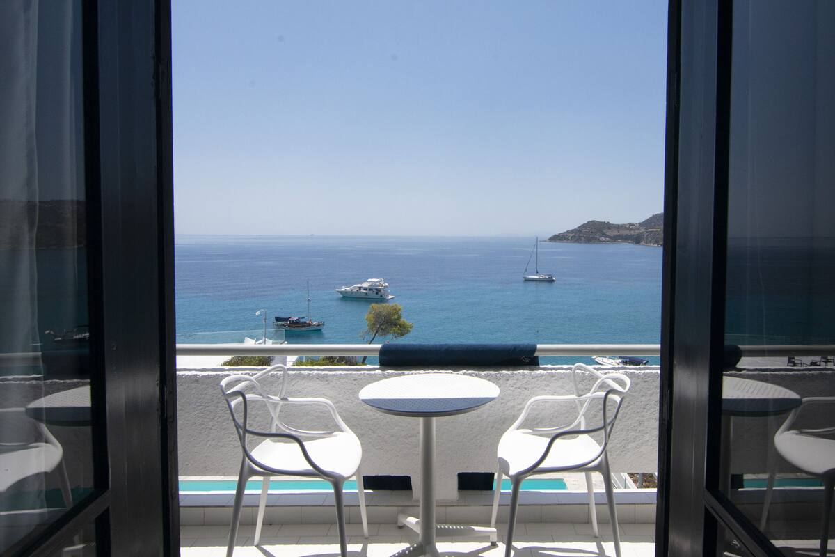 A balcony with white chairs and a small round table, overlooking a beach with boats and a clear blue sky.