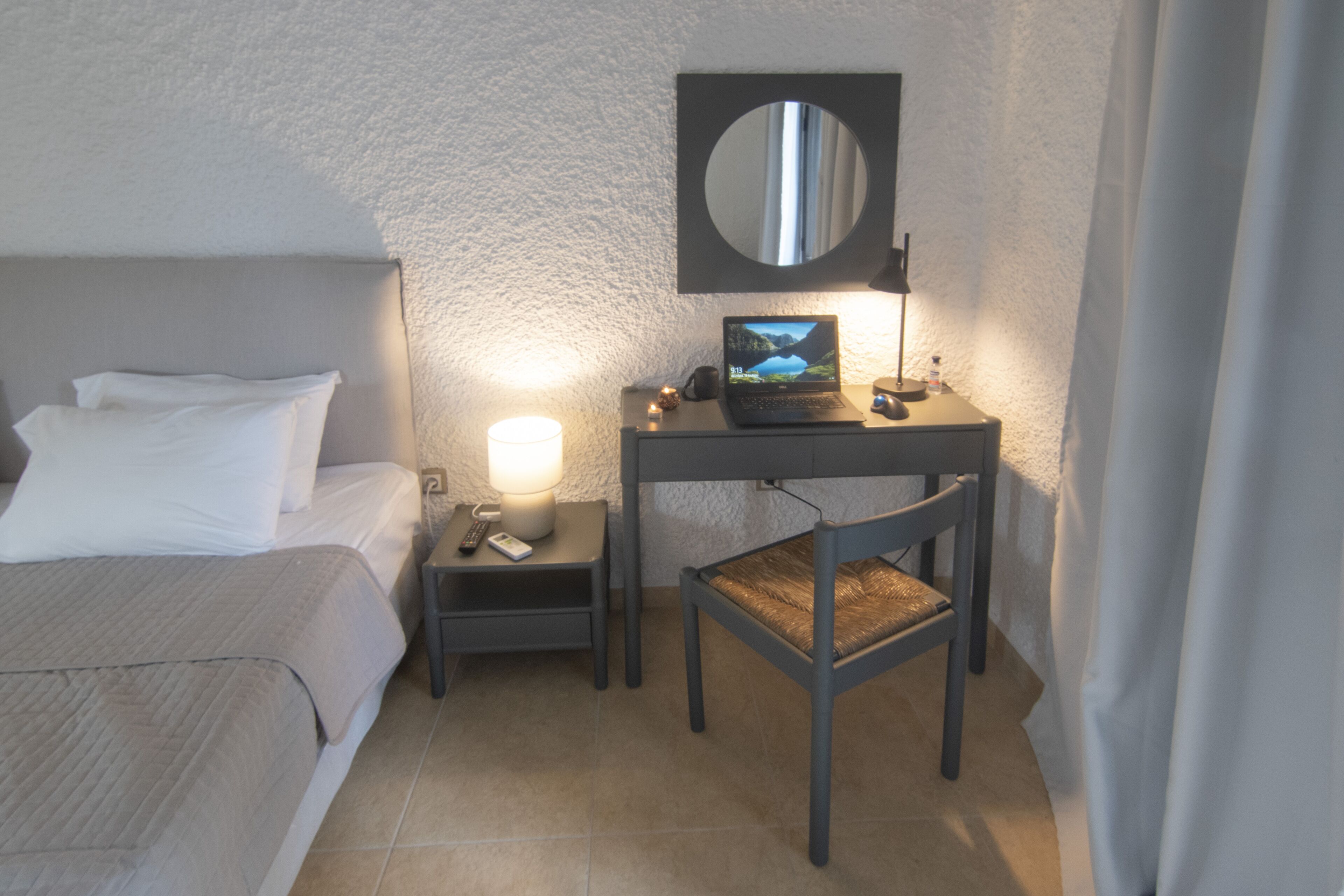 A hotel room with a bed, a desk with a laptop, a chair, a nightstand with a lamp, and a mirror on the wall.