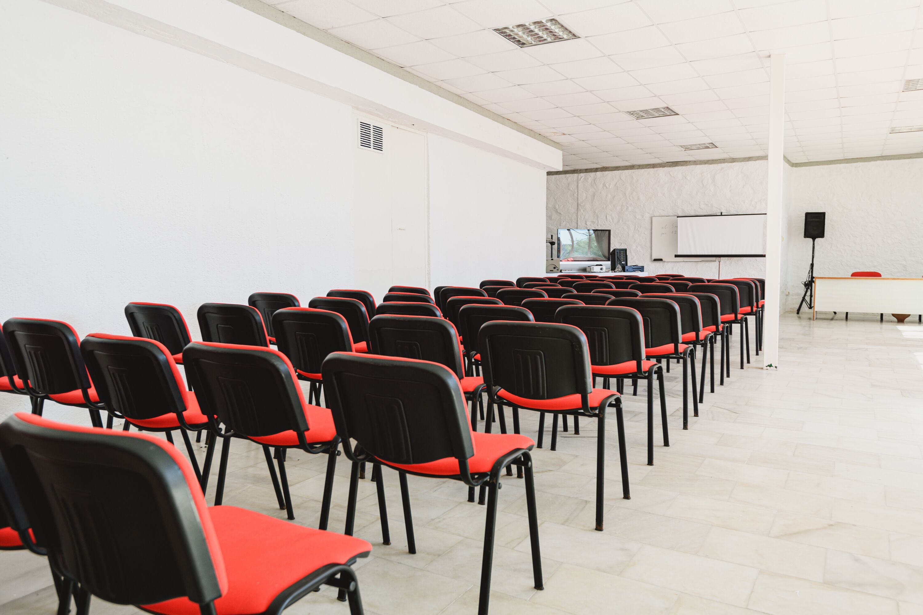 Un salón de conferencias con filas de sillas negras con cojines rojos, una pizarra, un proyector y un altavoz sobre un atril.