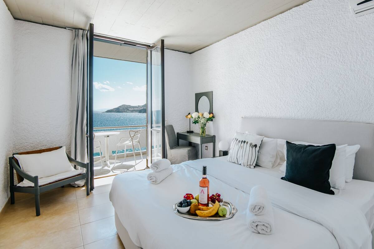 A hotel room with a bed, a small desk, a chair, and a balcony with a view of the sea.