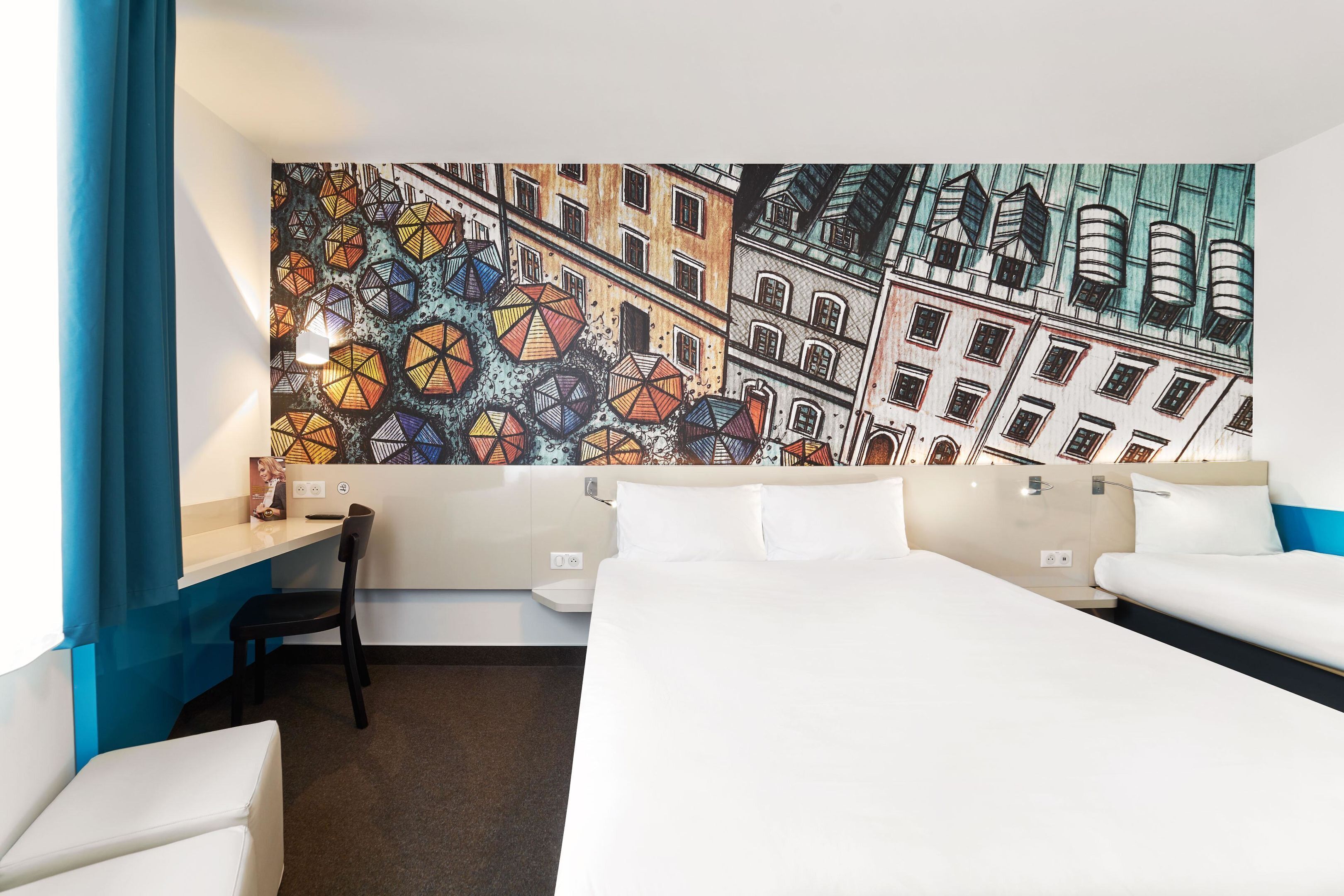 A hotel room with a large wall mural, a desk, two beds, and a chair.